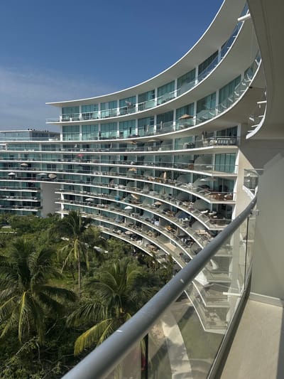 A modern high-rise building with a curved facade and balconies stretches upwards, partially obscuring a blue sky on the left. Each balcony is lined with chairs, tables, and umbrellas. Lush green palm trees and dense foliage fill the bottom right corner, contrasting with the building's sleek architecture. A gray metal pole runs diagonally across the upper right portion of the image, obscuring some of the structure and landscape. The image appears to be taken from a high angle, emphasizing the building's height and curvature.