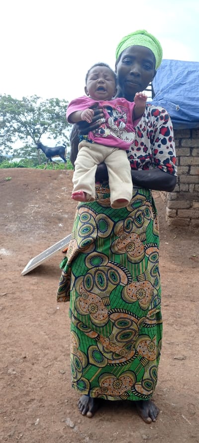 A woman stands barefoot on reddish dirt holding a crying baby wearing a pink Minnie Mouse shirt and beige pants. The woman wears a green patterned skirt, a patterned long-sleeved shirt, and a green head wrap. A solar panel lies on the ground beside her. In the background is a dirt mound, a black goat, and a tree. A structure with brick walls and a blue tarp covering is also visible.