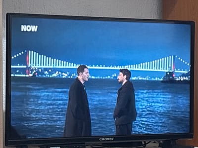 A television displaying a scene with two men standing opposite each other against a backdrop of a body of water and a bridge illuminated with lights. The word "NOW" is visible in the upper left corner of the screen. The television is a Crown brand. There is no indication of the location being Grohotno, Bulgaria, or any information to suggest a connection to the specified city.