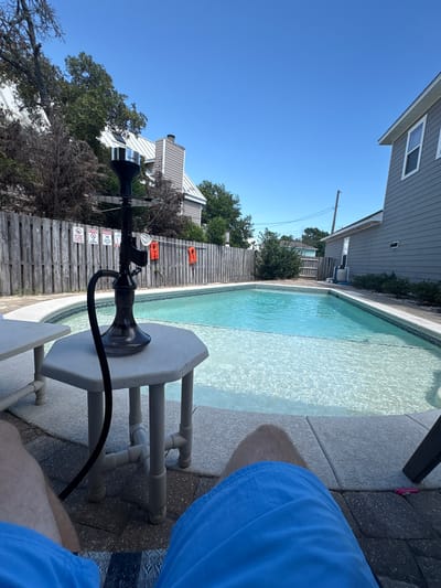 The image captures a sunny day in the backyard of a residential property, likely in Panama City Beach, Florida, United States. A person's legs, clad in blue shorts, are visible in the foreground, indicating they are seated, possibly on a lounge chair.
Dominating the scene is a swimming pool with crystal clear, turquoise water, reflecting the bright blue sky. The pool has a tiled edge and is surrounded by a light-colored patio.
On a small, octagonal, white plastic side table next to the pool sits a black hookah pipe. The hookah features a decorative stem and a dark-colored base, with a black hose extending from it.
In the background, a wooden privacy fence runs along the perimeter of the yard. Attached to the fence are several warning signs, including one that reads "NO GLASS CONTAINERS IN POOL AREA." There are also three bright orange life preservers hanging on the fence, suggesting a focus on water safety.
Behind the fence, a two-story house with gray siding and white trim is visible. The house has several windows, and a chimney can be seen on its side. Trees and other foliage are scattered throughout the scene, adding to the residential atmosphere. The clear blue sky indicates good weather.