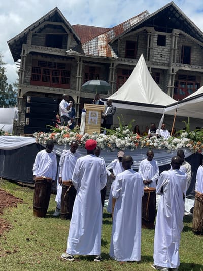 The image shows a large group of men in white robes gathered in front of a building in Rukoko, Democratic Republic of the Congo. The building appears to be under construction and has a concrete exterior. There is a stage set up in front of the building with a podium, flowers, and a tent. Several men are standing on the stage, one of whom is speaking. The men in the robes are playing drums and singing. The environment is green and grassy, with a dirt path leading away from the stage. The scene appears to be a celebration or a religious event.
