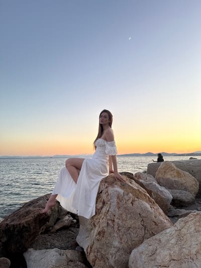 A young woman with long, dark hair sits on large rocks at the edge of the sea. She is wearing a white off-the-shoulder dress with a thigh-high slit, and her bare feet are visible. The sun is setting, casting a warm orange and yellow glow across the sky and water. In the distance, a faint crescent moon is visible in the pale blue sky. The rocky shoreline extends to the right of the woman, where another silhouetted figure can be seen sitting on the rocks. The sea is calm, with gentle waves lapping against the shore. The scene appears to be a pier or breakwater, characteristic of coastal areas like Paleo Faliro, Greece.
