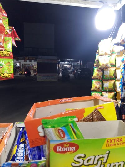 This is a medium-shot photograph taken at night, possibly on a street in Gampongpaya, Indonesia. The scene is illuminated by a bright light bulb hanging overhead, casting a strong glare and creating harsh shadows. In the foreground, several open cardboard boxes are filled with various snack items, including bags of chips and chocolate wafer bars.
One box prominently displays the brand "Roma" and "Sari" in large, bold lettering, suggesting these are popular local treats. Another box contains blue-wrapped snacks with the text "GORIORIO" visible. Hanging bags of chips, one with "JAGUNG" (corn) printed on it, are also visible in the upper left.
In the background, a dimly lit street scene unfolds with people and vehicles. A motorcycle is parked, and a few individuals are seen walking or standing near what appears to be a small shop or food stall. Signs with Indonesian text, such as "NASI GORENG BANDARA" (Bandara Fried Rice) and "AMRI STYLE," indicate local businesses. The overall atmosphere suggests a bustling night market or a roadside vendor at work. The lighting conditions contribute to a slightly mysterious or vibrant nocturnal ambiance.