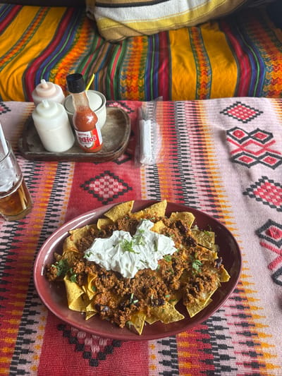 The image depicts a plate of nachos with ground meat, black beans, sour cream, and cilantro on a red plate placed on a colorful patterned tablecloth. Next to the plate, there is a wooden tray containing condiments, including a bottle of Frank's Red Hot sauce, two squeeze bottles with white sauce, and a small white container with a yellow straw. There's also a package of white napkins, a glass with a straw, and colorful pillow(s) on the left side of the image. The scene is photographed indoors. The location is Thong Sala, Thailand.