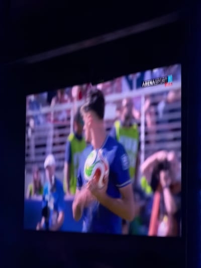 The image shows a close-up of a television screen displaying a live sports broadcast. The broadcast appears to be a soccer match, with a player in a blue jersey holding a soccer ball. The logo for "ARENASPORT 1" is visible in the top right corner of the screen. The background of the broadcast shows blurred images of spectators in a stadium. The television screen itself is framed by a dark border, suggesting it is set within a dimly lit room. The overall impression is that of someone watching a game on television. The prompt's request to include the location of Travnik, Bosnia and Herzegovina, cannot be fulfilled as there is no visual information in the image to suggest this specific location.