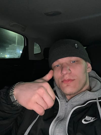 A young male subject with light skin and blue eyes is depicted inside the rear passenger compartment of a vehicle at night in Dobryanka, Russia. He wears a dark grey knitted beanie with a rectangular patch, a grey hooded sweatshirt, and a black track jacket featuring white trim and a white "NIKE" logo on the right chest. A metallic link bracelet is present on his left wrist. His left hand is raised, making a thumbs-up gesture. Through the vehicle's rear driver's side window, a brightly illuminated building with a visible "0+" sign is observed in the background. The vehicle's dark interior headliner and an overhead grab handle are also visible.