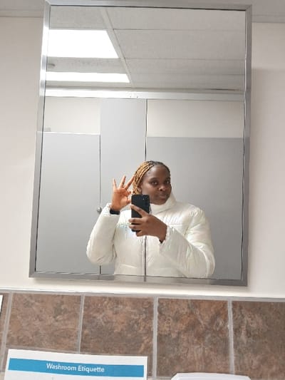 A person with blonde braids wears a white jacket and holds a smartphone, taking a mirror selfie in a bathroom. The mirror's frame is silver. The reflection includes parts of the bathroom, like a white wall and what appears to be a window with blinds. The area adjacent to the mirror shows brown floor tiles, and a sign is visible with the words "Washroom Etiquette" on it. The person's pose includes a hand gesture.