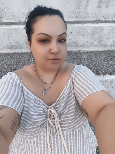 A woman with dark hair pulled back in a bun is taking a selfie outdoors. She is wearing a cream and beige striped off-the-shoulder top with a lace-up neckline and a cross pendant necklace. She has hoop earrings in her ears and tattoos on her arms. She is sitting on concrete steps, which appear to be part of a building's exterior. The background is a rough, grey concrete wall with horizontal lines. The scene is lit by natural daylight. The location appears to be an outdoor urban environment in Komotini, Greece.