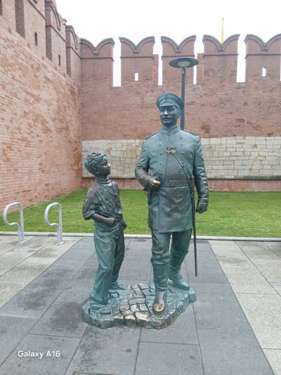 The image shows a bronze statue of a man in a military uniform standing next to a boy. The man is wearing a cap, a long coat, and boots, and he is holding a cane. The boy is wearing a shirt and pants. Both figures are looking in the same direction, towards the right of the frame. The statue is located in front of a brick wall with crenellations, which appears to be part of a fortress or historical building. There is a lamppost behind the man. To the left of the statue, there are two silver bicycle racks and a grassy area. The ground in front of the statue is paved with stone tiles. The image is taken during the daytime, with overcast sky. This is a statue located in Tula, Russia, commemorating Leo Tolstoy and a boy from his village.