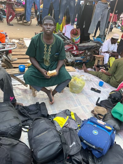 This image depicts a market scene in Nyanya, Nigeria. In the foreground, a young Black man is seated on a low wooden stool, wearing a green dashiki with gold embroidery. He is holding a piece of white bread in his hands. To his left, another person's legs are visible, wearing dark gray pants with a pink lightning bolt design.
The ground is covered with a patterned tarp, and a collection of bags, including large black duffel bags, a black Samsonite backpack, a blue child's backpack with space-themed decorations, and other assorted bags, are scattered around. To the left of the seated man, a blue plastic cup with some liquid and small objects inside is on a wooden plank. A red motorcycle is parked behind him.
In the background, the market is bustling with activity. Various stalls are visible, with clothing, particularly jeans, hanging from racks. People are seen browsing and interacting within the market environment. The overall scene suggests a vibrant, outdoor marketplace.