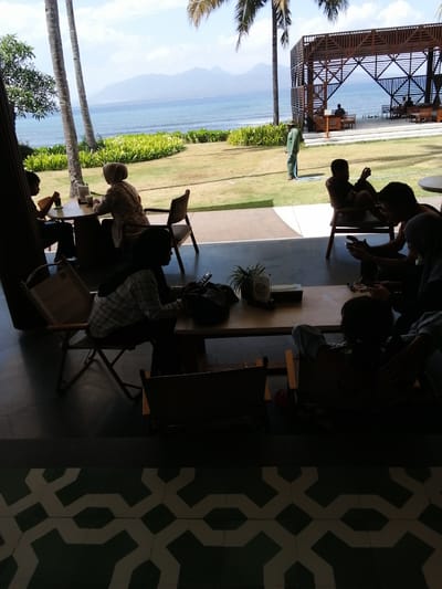 The image depicts a group of people enjoying a meal at an outdoor cafe overlooking a beach in Banyuwangi, Indonesia. The cafe features a wooden structure with seating areas, a bar, and a thatched roof. Palm trees are visible in the foreground, and the ocean stretches out to the horizon, with mountains in the distance. Several individuals are seated at tables, engaged in conversation and eating. The atmosphere appears relaxed and casual, with a focus on enjoying the scenic beauty of the location.