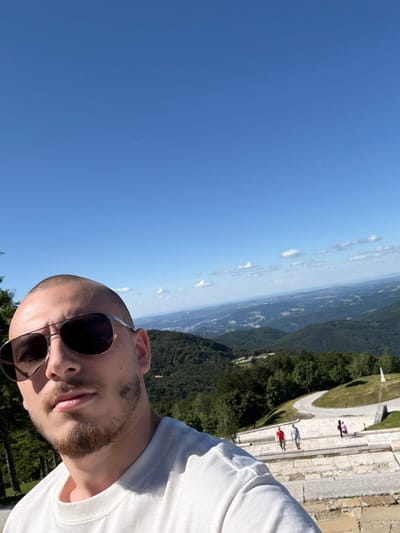 A man wearing sunglasses and a white t-shirt takes a selfie on a sunny day with a mountainous landscape in the background. The man is close to the camera, with his face occupying the lower left quadrant of the image. He has a shaved head and stubble. In the background, rolling green hills are covered in dense forest, with a winding road visible further up the slope. A few people can be seen on a stone walkway in the distance. The sky is a clear, bright blue with a few scattered white clouds. The location is the Shipka Pass in Bulgaria, a significant historical site.