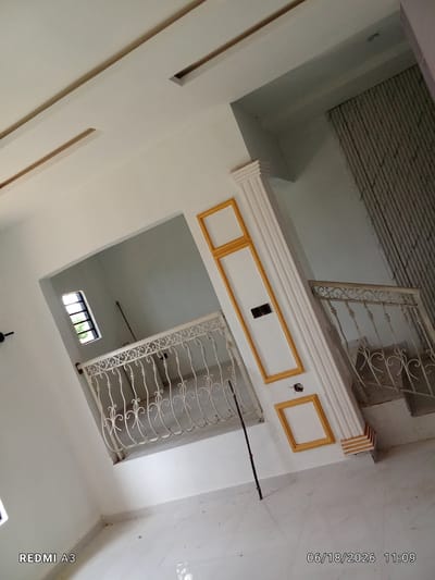 The image depicts an interior view of a building, likely a staircase or hallway. The walls are painted white and feature decorative elements, including recessed ceiling lights with gold trim and a fluted white column with gold trim accents. A white metal railing with ornate scrollwork lines a staircase on the right. On the left, a doorway opens into a room with a small window featuring horizontal bars. The floor appears to be tiled in a light color. A thin, dark stick or pole stands vertically near the column. The scene is brightly lit, suggesting daylight. The lower left corner displays the text "REDMI A3," and the lower right corner shows the date "06/18/2026" and time "11:09." The image quality is moderate, with some graininess. No people are visible in the frame. The location is identified as Nawfia, Nigeria.