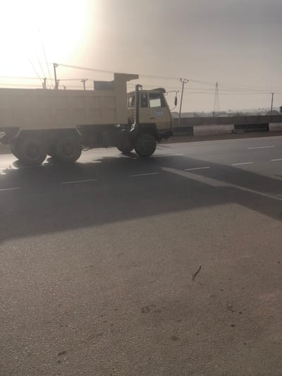 A dump truck sits on the roadside beside a paved road. Overhead power lines extend into the distance. The sky is overcast. Trees and vegetation are visible in the background near a roadside barrier. The road surface is asphalt.