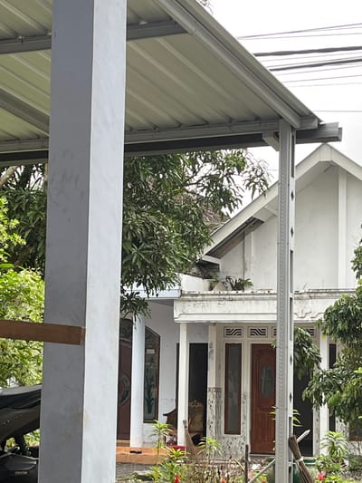 The image shows a tropical outdoor scene, possibly in Jambearum, Indonesia. The foreground is dominated by a light gray, corrugated metal roof structure supported by metal beams. Behind this structure, a white building with multiple stories is visible, featuring decorative patterned panels and what appear to be doors and windows. Several large trees with lush green foliage are present, some of which obscure parts of the building. The overall impression is of a residential or commercial property in a warm climate. There are no people visible, and the activity taking place is not discernible. Notable details include the architectural style of the building, the vibrant green of the trees, and the corrugated roofing material. The time of day and weather are not clearly indicated, but the bright lighting suggests daytime with possibly sunny conditions. No visible text is present in the image.