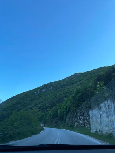 The image displays a picturesque mountainous landscape under a clear blue sky, viewed from what appears to be a vehicle. A steep, heavily vegetated mountain or hillside dominates the left and central parts of the frame, covered in dense green foliage.

On the right side, a paved road with a visible white line curves gently, running alongside the base of the mountain. Adjacent to the road, a concrete retaining wall, topped with a wire mesh system (likely for rockfall protection), is built into the hillside. Below the wall, exposed rocky ground can be seen, contrasting with the lush greenery. The sky, a vibrant, cloudless blue, occupies the upper portion of the image, indicating bright and sunny weather. A dark border on the far right suggests the photo was taken from inside a car or similar vehicle.

The scene conveys a sense of travel through a natural, mountainous environment, likely during the daytime given the bright illumination and clear sky. There are no people visible, and no discernible text. The setting aligns with a mountainous region such as the Stavroupoli Municipal Unit in Greece, featuring a road winding through natural terrain.