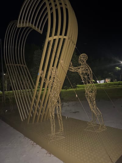 A wireframe sculpture of a heart, supported by vertical metal beams, is illuminated at night in Egorlykskaya, Russia. Two human-like wireframe figures are positioned in front of the heart sculpture. One figure stands on the left, facing the heart with outstretched hands. Another figure stands on the right, leaning against the heart with its hands on the structure. The ground beneath the sculpture is a metal surface with a diamond plate pattern, and in the background, trees and distant lights are visible.