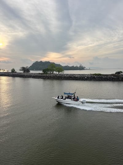 The image depicts a white motorboat moving rapidly through a body of water, likely a river or channel, in Pak Nam Chumphon, Thailand. The boat is leaving a large white wake behind it, indicating its speed. Several people are visible on board, their activities unclear but potentially related to fishing, given the fishing rods that are visible. A stone embankment lines the left side of the waterway, with trees growing along its edge. On the opposite side, the landscape features hills and trees silhouetted against a cloudy sky at sunrise or sunset. The sky is filled with light, streaky clouds, creating a diffused, atmospheric effect. The water is dark, reflecting the subdued lighting conditions.