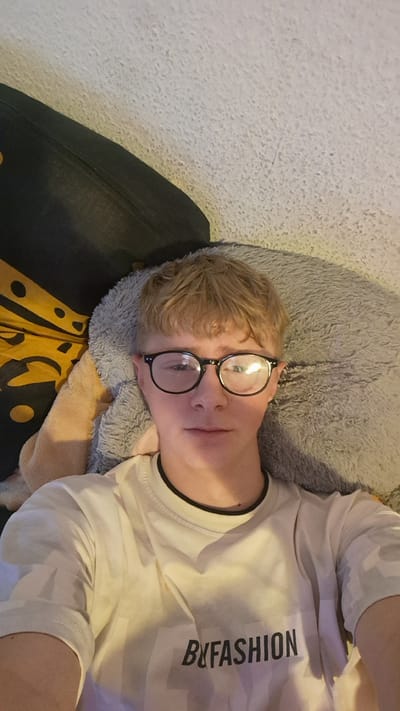 A person wearing a white t-shirt with black text that reads "B&FASHION" and round glasses is lying down. They are positioned on a fluffy grey pillow, with a black pillow and a fuzzy tan blanket visible in the background. The setting appears to be indoors, possibly a bedroom or living room, with a light-colored carpet on the floor. The lighting suggests it might be daytime or a well-lit indoor space. The person's expression is relaxed, and they are looking upwards towards the camera. The overall mood is casual and comfortable. No specific weather or time of day is discernible beyond the indoor context. The location context provided is Parchim, Germany, but there are no visual cues in the image to confirm this.