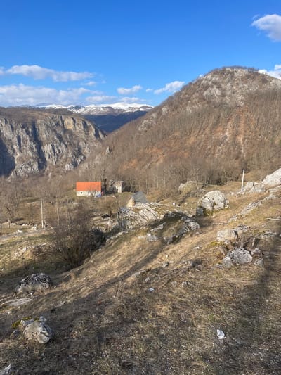 The image depicts a mountainous rural landscape in Mokro, Montenegro, under a clear blue sky with scattered white clouds. The scene is presented from an elevated viewpoint looking down into a valley.
In the immediate foreground, a dry, sloping terrain is visible, characterized by brown and pale green dry grass, exposed soil, and numerous light gray rocks and boulders. Sparse, leafless bushes and tree stumps are interspersed throughout this area. A piece of white crumpled debris is visible on the ground towards the lower right.
The midground features a small cluster of structures situated within a more level, partially vegetated area at the base of the slope. The most prominent building is a light-colored house with a distinct orange-red gabled roof. Adjacent to it on the right is another light-colored building, possibly a barn or an older dwelling, with a partially visible dark roof. To the right and slightly in front of these two buildings, a smaller, darker structure with a dark roof and lighter walls is present, appearing to be a shed or outbuilding. Utility poles with overhead lines are visible in the midground, with bare deciduous trees and bushes surrounding the structures and extending up the slopes to the right.
The background reveals a deep canyon or valley. On the left, a sheer, rocky cliff face rises dramatically. On the right, a densely forested mountain slope, primarily consisting of bare deciduous trees, ascends. In the distant background, a range of snow-capped mountains is visible beneath the horizon. There are no individuals or discernible actions occurring within the frame. The lighting suggests a bright, sunny day.
