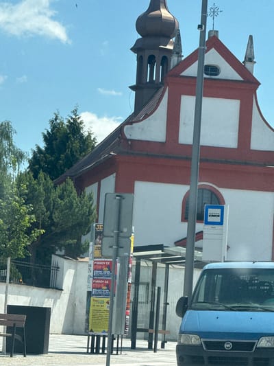 The image shows a street scene in Žďár nad Sázavou, Czechia. The focal point is a white and red building, possibly a church, with a domed tower. In front of the building, there is a bus stop shelter with a bus stop sign indicating "Horni". A blue van is parked in the foreground, partially obscuring the bus stop. Several posters are attached to a pole near the bus stop, some advertising concerts or events. Trees and a black trash bin are also visible. The sky is blue with some clouds.