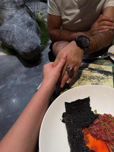 The image captures a dining scene, likely indoors, possibly in a restaurant. A man's arm, adorned with a wristwatch and bracelet, rests on the table. His hand is clasped by a woman's hand, distinguished by a manicure with a pink design. The table setting includes a plate with what appears to be a culinary dish consisting of raw minced meat, a black crispy element, and an orange sauce. Cutlery, including a fork and knife, is present. The table is covered with a patterned cloth. The floor appears to be a grey marble-like surface. There is a rock with some vegetation near the table. The image does not show Zhangjiajie.