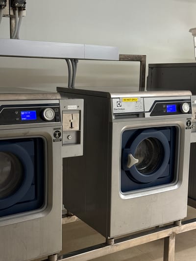 The image shows two commercial washing machines in a room. The washing machines are silver with blue doors and digital displays. One washing machine, on the left, has a digital display that reads "COLD 40", "5 MIN", and "EXTRACT". The other washing machine, on the right, has a yellow sticker that says "DO NOT OPEN" and the Electrolux Professional logo. Both washing machines have coin slots. The machines are situated on metal stands. Above the machines, there is a metal pipe and a light-colored wall. The floor appears to be tiled. The overall environment suggests a laundry room or a commercial establishment where laundry services are provided. There are no people visible in the image.