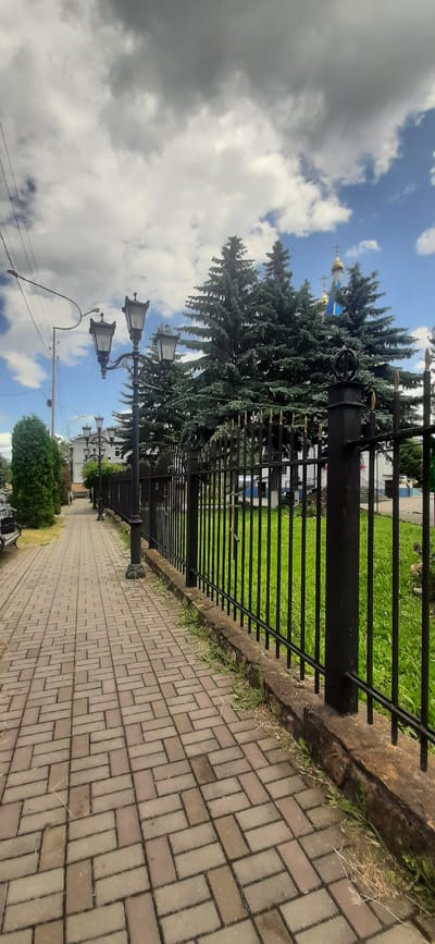 The image depicts a paved pedestrian walkway with brick-like pavers, extending into the distance on the left. A black metal fence with vertical bars and decorative finials runs along the right side of the walkway. Beyond the fence, a strip of bright green grass is visible, with some dry grass and weeds growing at the base of the fence. In the background, several tall, dark green evergreen trees are present, with the golden domes and blue roofs of a church visible behind them. The sky above is a mix of bright blue with scattered white and dark gray clouds. Several ornate black lampposts with multiple lamps are positioned along the walkway. The scene appears to be in a park or near a public building.
