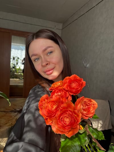 A young woman with long brown hair and light eyes holds a bouquet of bright orange roses close to her face. She is wearing a dark, silky top. The background includes a window, a plant, and what appears to be a bed. The lighting suggests it is daytime. There is no city of Dubrova in Russia, therefore, location cannot be confirmed.