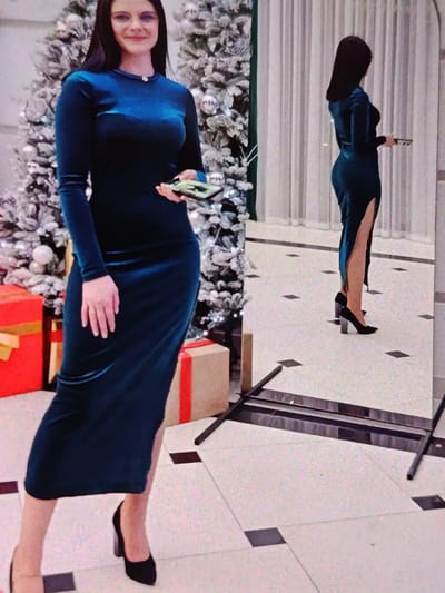 A woman stands in front of a decorated Christmas tree and a large mirror, holding a cell phone. She is wearing a long-sleeved, dark teal velvet dress with a high slit up the side and black high-heeled shoes. The Christmas tree is adorned with white and silver ornaments, and red gift-wrapped boxes are visible in the foreground. The woman is smiling, with long dark hair and glasses. The scene appears to be indoors, possibly a home or event space, with tiled flooring and curtains in the background. A reflection of the woman in the mirror shows her from the side, highlighting the dress's silhouette and the slit. The lighting suggests it could be daytime or evening. There is no visible text in the image.