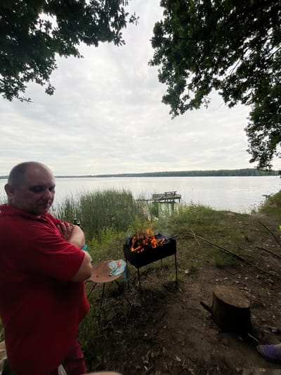 A man is lying on his back, wearing a red long-sleeved shirt, with his arms crossed. He is looking up at the sky. In front of him, a small campfire burns in a black grill. Behind the campfire, tall reeds grow along the shore of a lake. A wooden dock is visible in the water. The sky is overcast. The image appears to be taken outdoors, possibly at a campsite near a lake, based on the presence of the campfire and the natural surroundings. The scene seems relaxed and casual. The time of day is difficult to determine due to the overcast sky, but it is likely daytime.