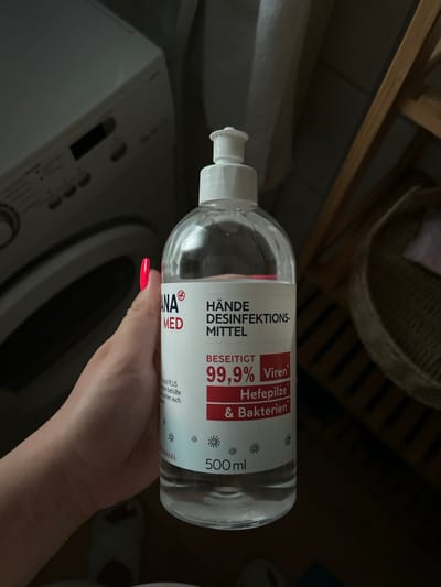 The image shows a close-up, vertically oriented view of a hand holding a clear plastic bottle of hand sanitizer. The hand has bright pink fingernails and is positioned in the foreground, obscuring part of the bottle and the background. The hand sanitizer bottle has a white pump dispenser and a label with German text. The label indicates that the product is a hand disinfectant that eliminates 99.9% of viruses, yeast fungi, and bacteria. It also specifies the volume as 500 ml and displays the brand "ANA MED". The background is dark and out of focus, but suggests an indoor setting. To the left of the bottle, the edge of a white appliance, possibly a washing machine, is visible. Above and behind the bottle, blurred wooden shelves and a woven basket can be seen, hinting at a home or storage area. The lighting is dim, and the overall impression is that the photo was taken indoors during the day or evening, without any specific indications of time of day or weather. The focus is on the product and the hand holding it.