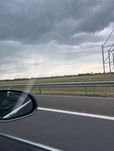 The image depicts a view from a car traveling on a highway. To the left of the road, a grassy field stretches towards the horizon under a cloudy sky. A guardrail runs parallel to the road, separating it from the field. The side mirror of the car is visible in the lower right corner. A white lane marking is present on the road. The location is near Balástya, Hungary, based on similar landscapes and infrastructure found in the area. There are no people visible. The scene is captured from a moving vehicle.