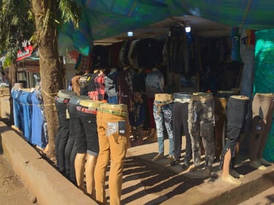 A clothing store in Lindi Street, Tanzania, is shown in this image. The store has a tarp-like canopy, supported by a series of metal poles. The store is located at the coordinates -3.355 latitude, 37.341 longitude. The shop is filled with mannequins wearing a variety of clothing, including jeans, pants, and shirts. The mannequins are all arranged in a row and stand on a concrete floor. Some of the mannequins have their bodies covered in a brown, flesh-toned substance, representing the color of human skin, while other mannequins are missing their legs. The mannequins are positioned at different heights, with some standing taller than others. Behind the row of mannequins is a rack of clothing with numerous items on it, including various colors and styles of jackets, shirts, jeans, and other pieces of clothing. The store is lit by overhead lights and is well-lit. The shop is set up on the side of a street. Behind the store, there is a large tree on the left side of the image. Behind the tree is a small structure covered in faded light blue paint with a yellow, orange, and white sign on the front of it. The sign has illegible writing on it. The store is situated in a bustling area of Lindi Street, Tanzania.