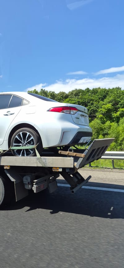 A white sedan is being transported on the back of a tow truck on a sunny day. The car is secured to the flatbed with green straps. The tow truck is driving on a paved road, with a guardrail and trees lining the right side of the road. The sky is blue with scattered white clouds. The scene appears to be on a highway outside of the city of Gorodechnoe, Russia.