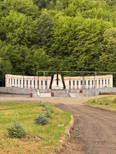 The image shows a monument with many plaques set against a backdrop of lush green trees. The monument appears to be made of concrete or stone and is structured in a way that suggests a place of remembrance. Rows of rectangular plaques are arranged in a grid pattern on the monument's facade. Some wooden benches are visible near the monument, providing seating. The ground around the monument is a mix of gravel paths, stone tiling, and grass. In the foreground, several small, young evergreen trees are planted in the grassy area. The sky is not visible, but the lighting suggests it is daytime, possibly overcast or with soft sunlight filtering through the leaves. The overall scene evokes a somber and reflective atmosphere, likely a memorial site. The location is identified as Dilijan, Armenia. There is no visible text in the image that can be read.