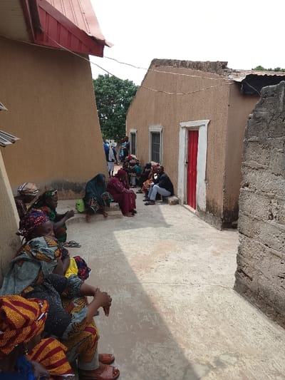 The image shows a group of people gathered in an outdoor space in a rural Nigerian settlement. There is a group of women sitting on the ground in the foreground, wearing traditional African dress and headscarves. The women are all looking in the same direction, towards a group of people standing and sitting in the background. There are two men and one woman in the background. The men are both wearing shirts and trousers. The woman is wearing a bright red dress. The background shows an exterior of a mud-brick building with a red door. The building is partially obscured by a large, green tree. The setting is the town of Mukaiya, Nigeria, with a latitude of 8.4 and a longitude of 8.343. The ground is covered with a mixture of sand and gravel. The walls of the buildings are made of mud bricks.