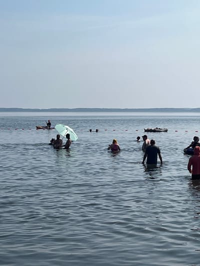 The image depicts a serene summer day at a beach in Surakovo, Russia. The scene is set on a wide, calm body of water, likely a lake or bay, with a hazy horizon where the water meets a faint shoreline under a clear, pale blue sky. Numerous individuals are engaged in various water activities. In the foreground, several people are wading in the water. To the left, two individuals stand under a light blue umbrella, partially submerged. Further back, a person is paddling a canoe, and another is lying on an inflatable raft. Several other people are scattered throughout the water, some swimming, others standing. A line of buoys demarcates a section of the water. The overall atmosphere is one of leisurely enjoyment on a warm day.