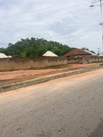 This is a landscape photograph taken from the side of a paved road in Minda, Nigeria. The road appears to be in fair condition with some visible patches and stains. A concrete curb separates the road from a dirt path, which runs alongside a brick wall. Beyond the wall, the terrain slopes upwards, covered in lush green vegetation and trees. The sky is overcast with scattered clouds, suggesting it might be a cloudy day.
In the background, several buildings with corrugated iron roofs are visible, nestled against the hillside. There are also power lines stretching across the frame, indicating the presence of infrastructure. The overall scene depicts a rural or semi-urban environment in Nigeria, characterized by a mix of natural landscape and man-made structures. There is no visible text in the image, nor any clear indication of a specific activity or event taking place, other than it being a typical day in this location. The lighting suggests it is daytime, but the overcast sky limits strong shadows, creating a diffused light. The image evokes a sense of everyday life in a Nigerian setting, with the natural environment playing a prominent role.