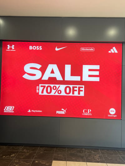 The image is a photograph of a large digital advertisement display in Newry, United Kingdom. The advertisement is primarily red and features the word "SALE" prominently in large white lettering. Below "SALE," it states "UPTO 70% OFF" within a white rectangle. The advertisement also showcases logos of various brands including Under Armour, BOSS, Nike, Nintendo, Adidas, Skechers, PlayStation, Puma, C.P. Company, and Slazenger. The display is mounted on a dark gray wall panel system, with a tiled floor visible at the bottom of the image. There are no people or animals present in the image.