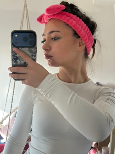 Location: Bouxwiller, France.
Scene: An indoor setting depicts a female individual taking a self-portrait in a mirror. The subject has dark, curly hair contained by a bright pink, plush headband featuring two prominent ear-like structures at the top. She is wearing a form-fitting, long-sleeved, off-white top and a thin gold-colored necklace. Her right hand is extended, holding a dark-colored smartphone, identifiable as an iPhone due to its triple camera array and dark green/black protective case. The phone case displays "PO COOL" vertically on its left side and a pattern of martini glasses, an 8-ball, stars, and leopard print bows on its rear surface. The individual's head is tilted slightly to her right, with her gaze directed past the phone, observing her reflection. The background is out of focus, revealing a partial view of a light-colored woven hanging chair on the left. Illumination is bright and even throughout the scene.