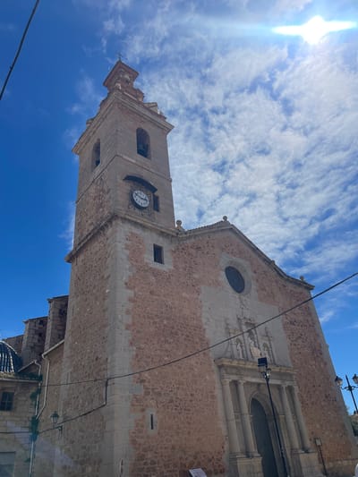 The image shows the upward view of the Church of Saints John in Puçol, Spain. The architecture is predominantly stone and brick, with a clock tower visible. The sky is partly cloudy, and the sun is shining brightly. The perspective emphasizes the height of the church. The building's façade shows signs of age and weathering. Electrical wires are visible against the sky and building.