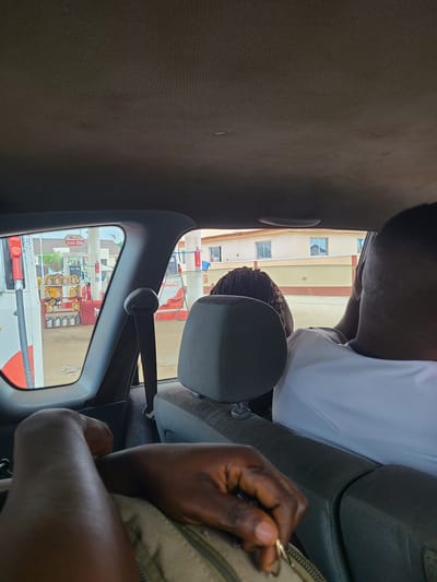 The image is taken from inside a vehicle, looking out the passenger window. The vehicle is at a gas station in Karu, Nigeria. In the foreground, a person's arm and hand are visible, resting on their lap. Further back, another person's head and shoulder are seen, also inside the vehicle. Through the window, rows of engine oil containers are displayed on shelves at the gas station. The gas pumps themselves are also visible, along with buildings in the background. The ceiling of the vehicle is visible at the top of the frame.