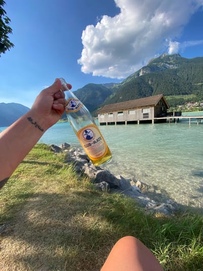 A person's arm, featuring a tattoo on the wrist, is holding a glass bottle of Club-Mate beverage. The bottle's label is visible, displaying the brand name and text in both German and English, mentioning "Original" and "Refreshing drink based on mate tea" with a note on caffeine content. The arm is positioned in the foreground, with the bottle extending towards a scenic background.

The scene is set by a clear, shallow body of water, likely a lake, with a wooden structure built on stilts extending into the water. The water is crystal clear, allowing the sandy bottom and scattered rocks to be seen. Beyond the water, lush green mountains rise steeply, covered in dense forest. The sky is a vibrant blue with scattered white clouds, suggesting a sunny day. A grassy area with some dry patches and rocks borders the water. The overall impression is one of a relaxed outdoor setting, possibly a lakeside leisure area, with a focus on enjoying a refreshing drink amidst nature. The quality of light suggests it is daytime, likely late afternoon given the warmth of the light and shadows. The text on the bottle is "CLUB-MATE Original Erfrischungsgetränk auf Mate-Tee-Basis mit Koffeingehalt" and "Listen to your heart" in Arabic.