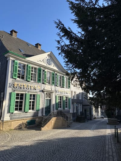 The photograph depicts the Deutsches Röntgen-Museum in Remscheid, Germany. The museum building is characterized by a white facade with multiple windows and green shutters. The building has a gable roof with chimneys, and a round window is visible near the roofline. The sign "Röntgenmuseum" is vertically displayed on the building facade. A large tree partially obscures the upper portion of the building and sky. The street beside the building is paved with cobblestones. The sky is blue.