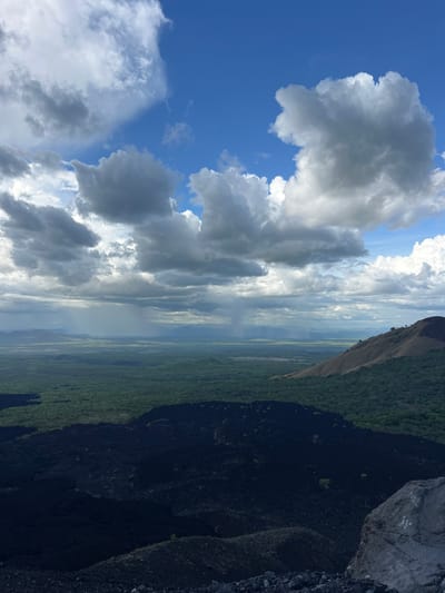 An eye-level, wide-angle, vertical shot depicts a vast, dramatic landscape dominated by dark volcanic rock in the foreground and a lush green expanse stretching to the horizon under a dynamic sky. The immediate foreground is a rugged, uneven terrain composed of dark, porous volcanic rock, with a few scattered grey rocks and a larger, rough-textured grey boulder on the right edge of the frame. Beyond this, a dense, dark green forest covers a gently sloping area, transitioning into a flatter, lighter green plain that extends to distant, hazy blue mountains. A patch of rain is visible as a grey, misty curtain in the middle ground, suggesting a weather event in progress. The sky above is a mix of deep blue and large, billowing grey and white clouds, with some areas of brighter white suggesting sunlight breaking through. The overall impression is one of raw, natural beauty and geological activity, possibly from an elevated vantage point overlooking a volcanic crater or lava field. The location is identified as Masaya Volcano National Park, Nicaragua.