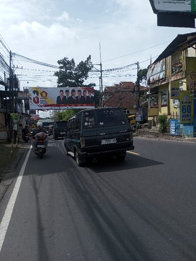 The image shows a street scene with a black van and a motorbike in the foreground. A large political banner stretches over the road, displaying images of several men in suits. Buildings line the street, with a laundry sign advertising a 60-minute service. Utility poles and wires are visible above. The license plate on the van reads "F 1532 HX". Signs with the text "50" and "60" are apparent.