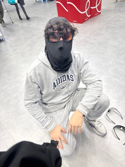 A person wearing a grey Adidas tracksuit, black balaclava, and orange tinted sunglasses sits cross-legged on a light grey floor. White sneakers and a few pairs of sunglasses rest near them. A few red boxes are in the background. Another person wearing a black coat is also in the image.