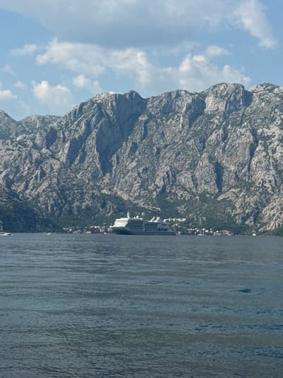 The image depicts a cruise ship docked near the town of Prčanj, Montenegro, situated along the Bay of Kotor. The ship is positioned close to the shore, with a backdrop of steep, rocky mountains covered in vegetation and partially obscured by low clouds. The water is calm, with visible ripples and reflections. The sky is blue with scattered white clouds. Buildings and other urban structures are faintly visible on the shoreline between the ship and the mountain.