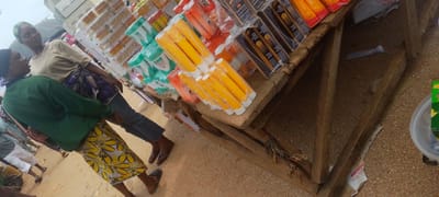 In Baissa, Nigeria, a market scene unfolds with a close-up view of two women interacting near a wooden stall laden with various consumer goods. The woman on the left, wearing a green long-sleeved shirt and a yellow and white patterned skirt, is positioned in the foreground. Her dark hair is covered with a headscarf. To her right stands another woman, wearing a light brown t-shirt and a patterned blue and white skirt, her head also covered with a headscarf. They appear to be engaged in a conversation, with their bodies oriented towards each other.

The stall, constructed from rough wooden planks, displays an assortment of products, predominantly cosmetics and toiletries, neatly packaged and stacked. Prominent among these are rows of yellow cylindrical tubes, some wrapped in plastic, and orange boxes with visible branding such as "Frya." Other items include green packaged products and dark boxes with text indicating "Intensive Toning" and "Solaray." The ground surface is composed of gravel and sand, typical of an outdoor market environment. The overall lighting suggests an overcast day. The image captures a candid moment within a bustling commercial setting, highlighting everyday life in Baissa.